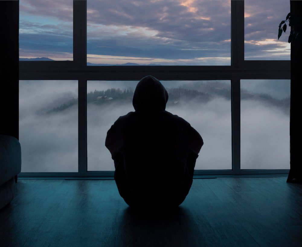Person mit Kapuze sitzt in der Dämmerung vor einem großen Fenster mit Blick auf Nebel und Berge