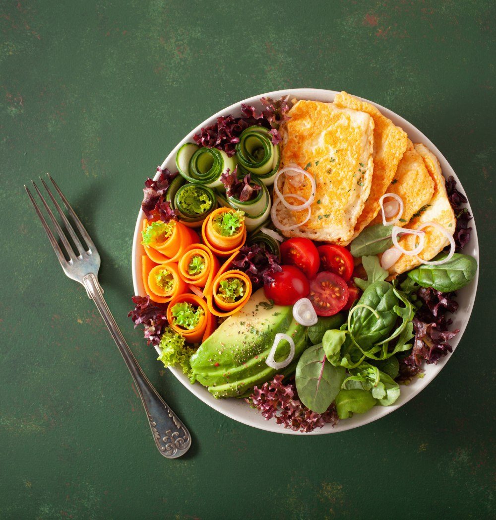 Bunte Bowl mit Halloumi, Avocado, Tomaten, Spinat und Gemüseröllchen auf grünem Untergrund, daneben eine Gabel